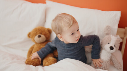 Adorable blond toddler sitting on bed with teddy bear at bedroom
