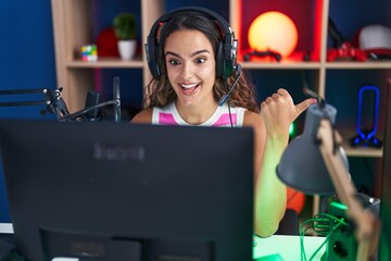 Young hispanic woman playing video games pointing thumb up to the side smiling happy with open mouth