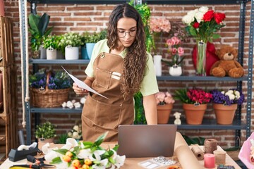Young beautiful hispanic woman florist using laptop reading document at flower shop