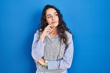 Young brunette woman standing over blue background with hand on chin thinking about question, pensive expression. smiling with thoughtful face. doubt concept.