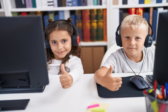 Adorable boy and girl students using computer and headphones doing thumbs up gesture at classroom