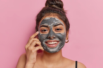 Portrait of good looking cheerful woman applies cosmetic clay mask smiles toothily looks happily at camera stands bare shoulders isolated over pink background enjoys softness. Cosmetology treatment