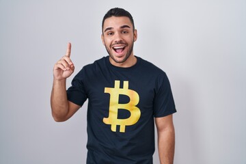 Young hispanic man wearing bitcoin t shirt pointing finger up with successful idea. exited and happy. number one.