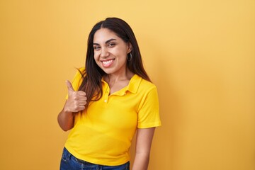 Young arab woman standing over yellow background doing happy thumbs up gesture with hand. approving expression looking at the camera showing success.