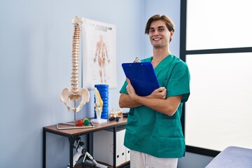 Young caucasian man pysiotherapist smiling confident holding clipboard at rehab clinic