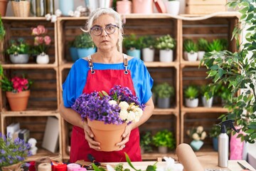 Middle age woman with grey hair working at florist shop holding plant skeptic and nervous, frowning...