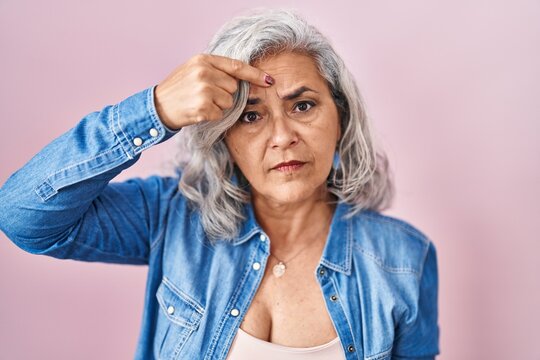 Middle age woman with grey hair standing over pink background pointing unhappy to pimple on forehead, ugly infection of blackhead. acne and skin problem