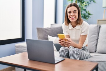Young beautiful hispanic woman using smartphone and laptop at home