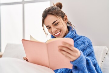 Young beautiful hispanic woman reading book sitting on bed at bedroom