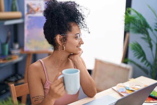Young hispanic woman artist using laptop drinking coffee at art studio