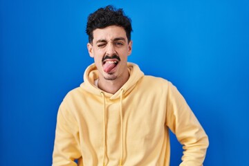 Hispanic man standing over blue background sticking tongue out happy with funny expression. emotion concept.