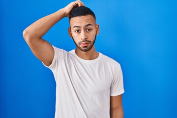 Young hispanic man standing over blue background confuse and wondering about question. uncertain with doubt, thinking with hand on head. pensive concept.