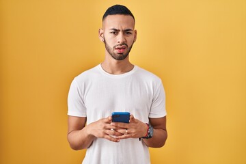 Young hispanic man using smartphone typing message clueless and confused expression. doubt concept.