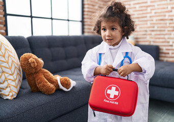 Adorable hispanic girl wearing doctor uniform holding first aid kit box at home