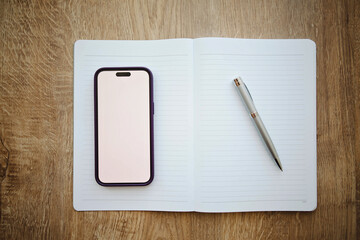 Overhead view of a Mobile phone with a blank white screen and a pen on an open notebook