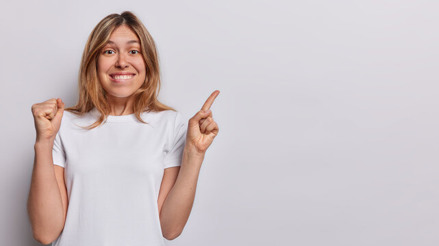 Horizontal shot of cheerful woman clenches fist and bites lips points index finger at copy space celebrates positive news dressed in casual t shirt isolated over white background. Wow great offer