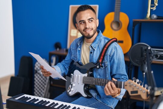 Young hispanic man musician playing electrical guitar reading music sheet at music studio