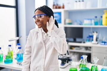 African american woman working at scientist laboratory smiling with hand over ear listening and hearing to rumor or gossip. deafness concept.
