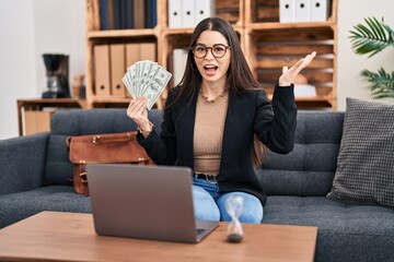 Young woman working at consultation office holding money celebrating victory with happy smile and winner expression with raised hands