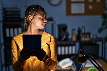 African american woman with braids working at the office at night with tablet looking to side, relax profile pose with natural face and confident smile.