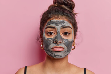 Portrait of displeased dark haired woman looks disappointed has sad facial expression applies clay nourishing mask for reducing fine lines poses bare shoulders isolated over pink background.