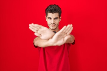 Young hispanic man standing over red background rejection expression crossing arms and palms doing...