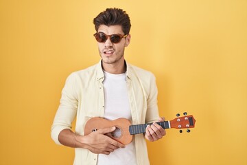 Young hispanic man playing ukulele over yellow background clueless and confused expression. doubt...
