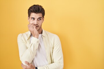 Young hispanic man standing over yellow background looking stressed and nervous with hands on mouth...