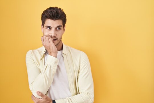 Young hispanic man standing over yellow background looking stressed and nervous with hands on mouth biting nails. anxiety problem.