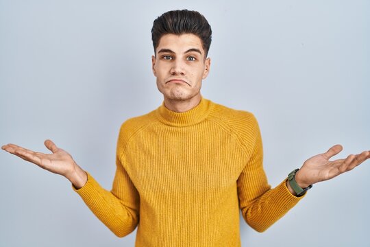 Young hispanic man standing over blue background clueless and confused expression with arms and hands raised. doubt concept.