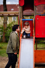 little toddler boy is going to slide down the hill in spring. Dad in a green sports jacket looks at the child.