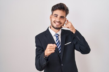 Young hispanic man with tattoos wearing business suit and tie smiling doing talking on the telephone gesture and pointing to you. call me.