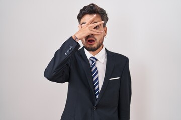 Young hispanic man with tattoos wearing business suit and tie peeking in shock covering face and eyes with hand, looking through fingers with embarrassed expression.