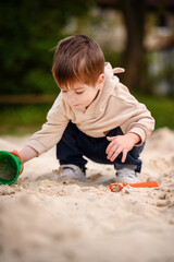 Little toddler boy sits in the sandbox and plays with toys in hoodie and jeans
