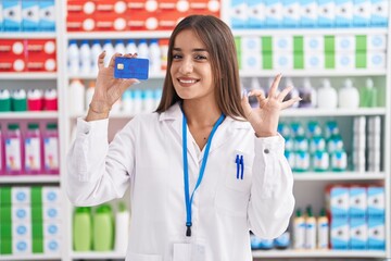 Young brunette woman working at pharmacy drugstore holding credit card doing ok sign with fingers, smiling friendly gesturing excellent symbol