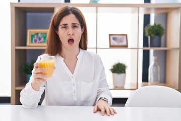 Brunette woman drinking glass of orange juice in shock face, looking skeptical and sarcastic, surprised with open mouth