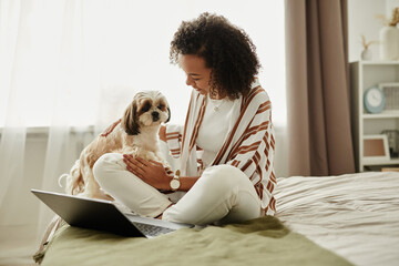 Candid portrait of young black girl relaxing at home sitting on bed with cute pet dog and using laptop, copy space