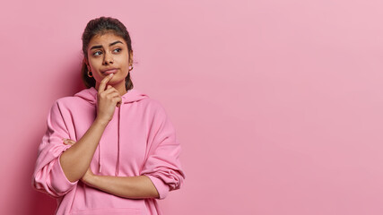 Studio shot of pretty thoughtful Indian woman holds chin concentrated aside being deep in thoughts...
