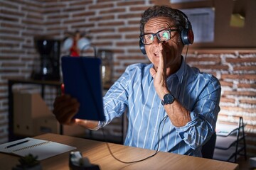 Middle age hispanic man using touchpad sitting on the table at night hand on mouth telling secret rumor, whispering malicious talk conversation