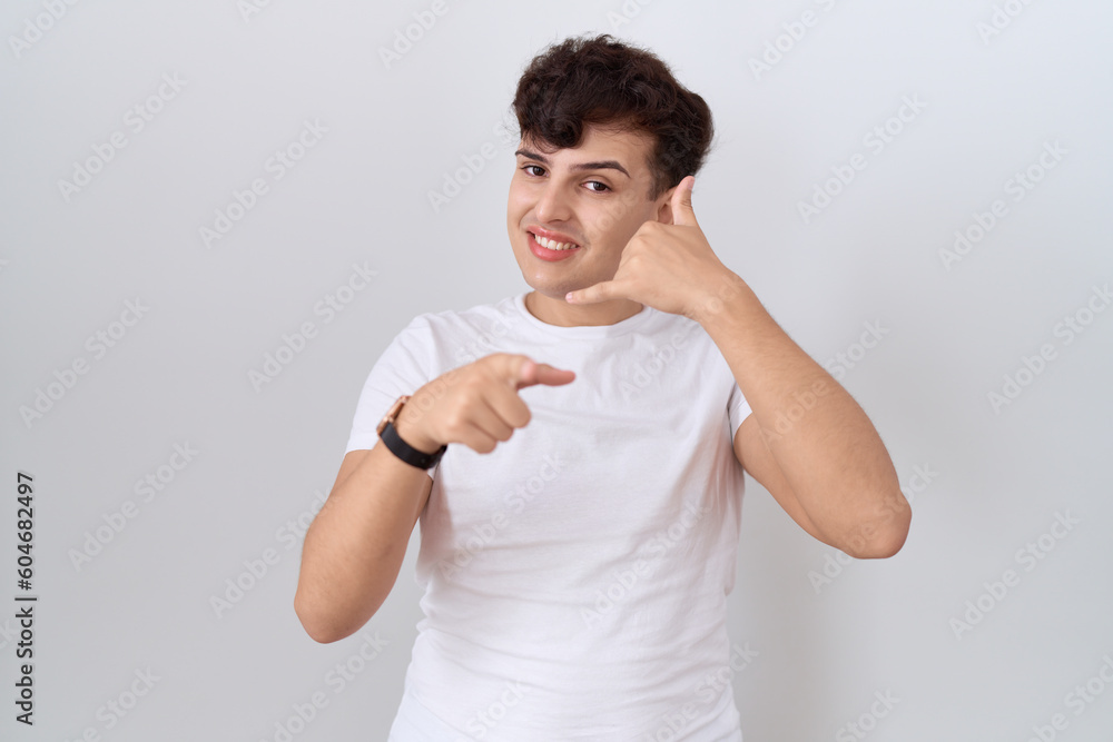 Wall mural Young non binary man wearing casual white t shirt smiling doing talking on the telephone gesture and pointing to you. call me.