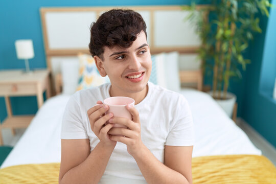 Non binary man drinking cup of coffee sitting on bed at bedroom