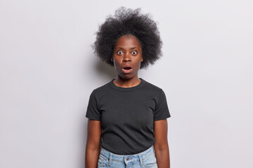 Frightened Afro woman with dark bushy hair whitnesses crime stares with shocked scared expression keeps mouth opened dressed in black t shirt jeans isolated over white background. No it cant be true - obrazy, fototapety, plakaty