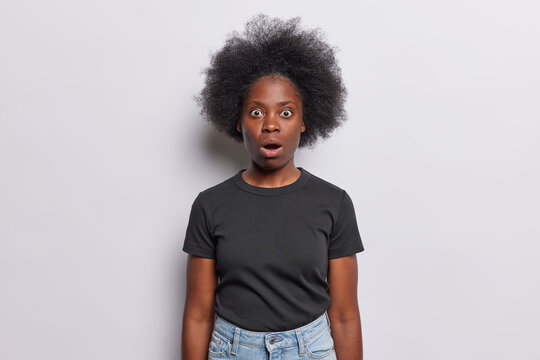 Frightened Afro woman with dark bushy hair whitnesses crime stares with shocked scared expression keeps mouth opened dressed in black t shirt jeans isolated over white background. No it cant be true