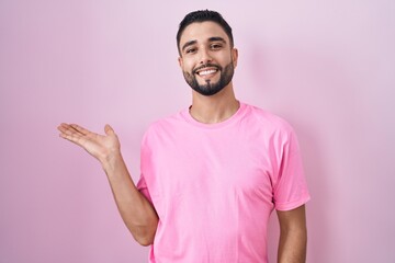 Hispanic young man standing over pink background smiling cheerful presenting and pointing with palm of hand looking at the camera.