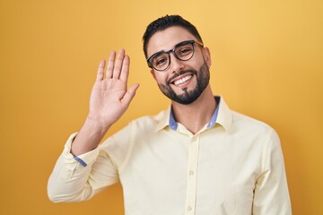 Hispanic young man wearing business clothes and glasses waiving saying hello happy and smiling, friendly welcome gesture