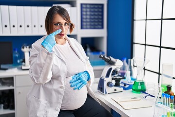 Pregnant woman working at scientist laboratory pointing with hand finger to face and nose, smiling cheerful. beauty concept
