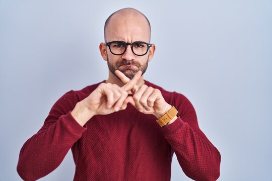 Young bald man with beard standing over white background wearing glasses rejection expression crossing fingers doing negative sign