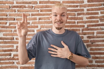 Young caucasian man standing over bricks wall smiling swearing with hand on chest and fingers up,...
