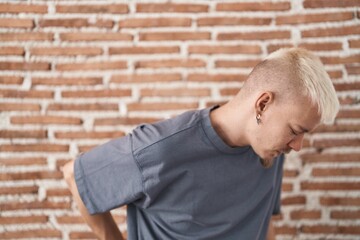Young caucasian man standing over bricks wall suffering of backache, touching back with hand, muscular pain