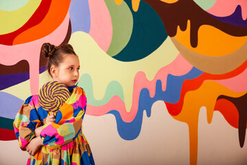 Pensive little girl posing with round large lollipop at multi colours wall backdrop, thought looking away. Lovely kid in colorful dress holding candy on stick. Summer sweet concept. Copy ad text space